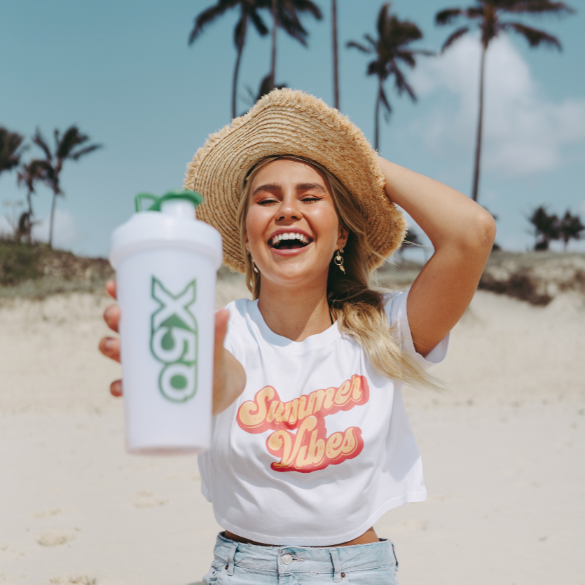 Smiling woman in a "Summer Vibes" shirt and straw hat on the beach, holding an X50 shaker bottle, promoting summer energy.